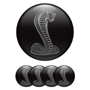 May include: A set of five circular emblems featuring a glossy black background with a centered silver-toned cobra snake graphic. The cobra is depicted in a coiled, upright position with a textured scale pattern and an open mouth, rendered in a metallic finish.