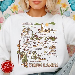 May include: White sweatshirt featuring a colorful map of the Pride Lands from The Lion King. The map includes Pride Rock, Elephant Graveyard, and Rafiki's Tree. The text "The Lion King" and "Pride Lands" are also visible.