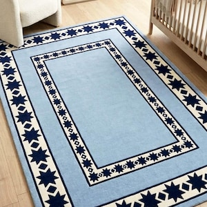 Puede incluir: Una alfombra rectangular con un campo central azul claro rodeado por un borde de color crema. El borde está decorado con un patrón repetitivo de estrellas azul marino de ocho puntas y pequeñas formas de diamante en un diseño geométrico.