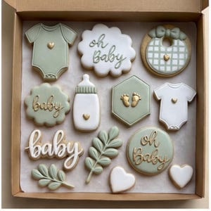 Puede incluir: Una caja de galletas con temática de bebé, que incluye bodies, baberos y biberones. Las galletas están decoradas en blanco y verde salvia con detalles dorados. Algunas galletas tienen las palabras "oh baby" y "baby".