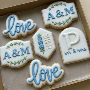 May include: Box of decorated sugar cookies with white icing and blue lettering. Cookies include the word "love", initials "A&M", the date "06 25 22", and "mr. & mrs.". Some cookies have floral accents.