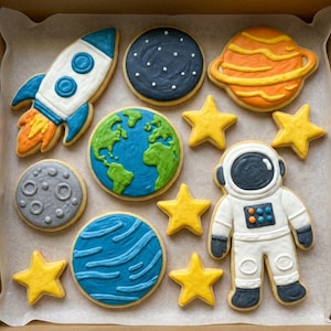 Può includere: Una scatola di biscotti decorati a tema spaziale. I biscotti includono un razzo, pianeti, stelle, un astronauta, la Terra e la luna. I biscotti sono decorati con glassa blu, arancione, gialla e grigia.