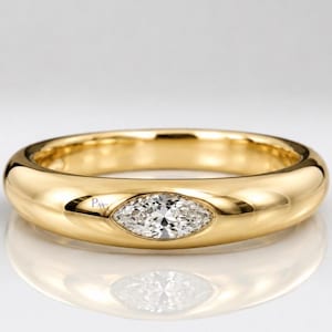 May include: A polished yellow gold band ring featuring a single marquise-cut clear gemstone set flush into the centre. The metal surface is smooth and rounded, with the letters PAG engraved in small, dark font near the gemstone setting.