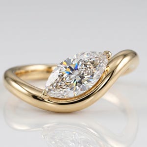 Op de afbeelding: Een gouden verlovingsring met een marquise-geslepen diamant. De diamant is gezet in een bezel-zetting en de band loopt rond de vinger. De ring is hoogglans gepolijst en reflecteert licht.