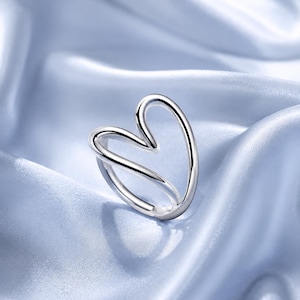 May include: A polished silver-tone ring featuring an open, continuous wire design that forms a stylized heart shape. The metal has a smooth, reflective surface, and the band curves to create the heart outline with a single, fluid metal loop.