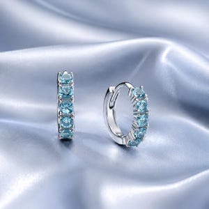 May include: A pair of silver hoop earrings featuring a row of five round-cut light blue gemstones set along the front of each hoop. The earrings have a polished metallic finish and a hinged closure mechanism for secure wear on the earlobe.