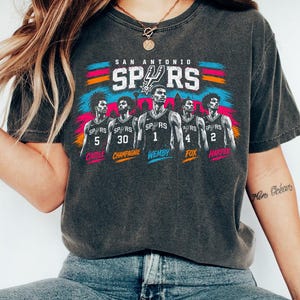 May include: A charcoal gray t-shirt featuring a graphic of five San Antonio Spurs basketball players in jerseys numbered 5, 30, 1, 4, and 2. The names Castle, Champagnie, Wemby, Fox, and Harper are printed below each player with colorful brush strokes.