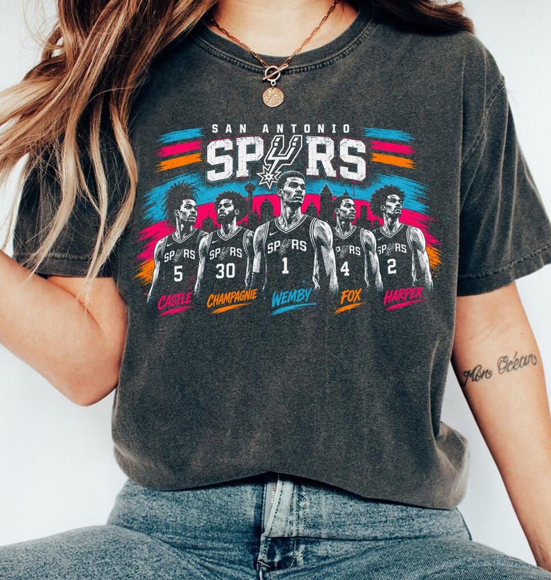 May include: A charcoal gray t-shirt featuring a graphic of five San Antonio Spurs basketball players in jerseys numbered 5, 30, 1, 4, and 2. The names Castle, Champagnie, Wemby, Fox, and Harper are printed below each player with colorful brush strokes.