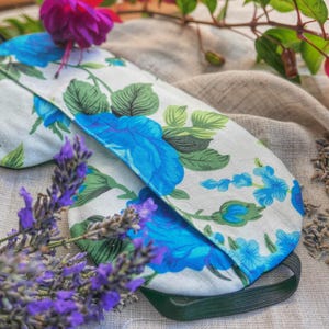 May include: A sleep mask featuring a white fabric base with a vibrant blue rose and green leaf floral pattern. The mask includes a black elastic strap and is surrounded by purple lavender sprigs, pink fuchsia flowers, and dried lavender buds.