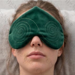 May include: A dark green eye mask with a spiral stitched design. The mask is made of a soft material and is designed to cover the eyes. The mask is being worn by a person.