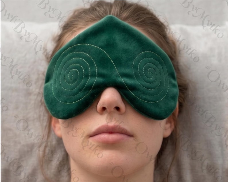 May include: A dark green eye mask with a spiral stitched design. The mask is made of a soft material and is designed to cover the eyes. The mask is being worn by a person.