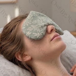 May include: A light green eye mask with a white leaf pattern covers the eyes of a person lying down. The mask is made of soft fabric and appears to be filled with a soothing material. The person is resting on a gray pillow.