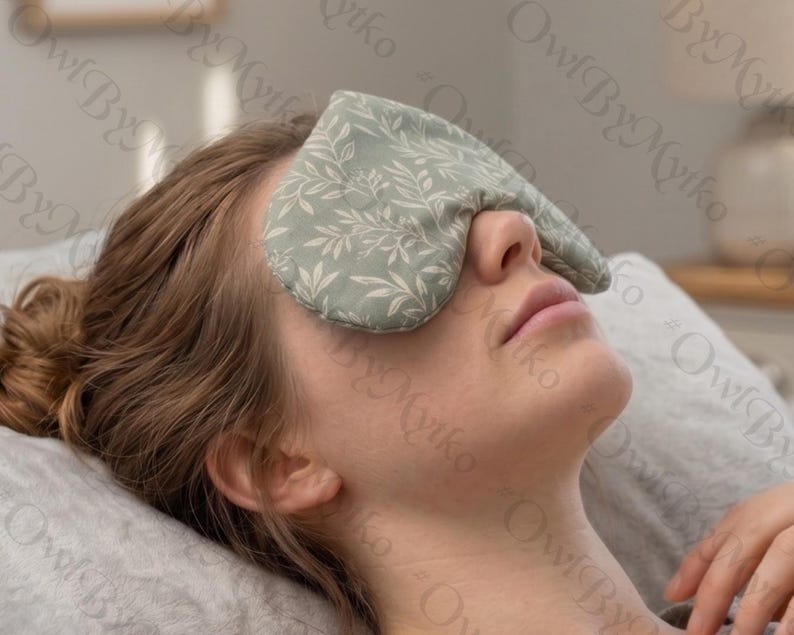 May include: A light green eye mask with a white leaf pattern covers the eyes of a person lying down. The mask is made of soft fabric and appears to be filled with a soothing material. The person is resting on a gray pillow.