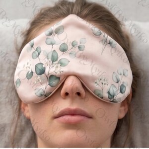 May include: A light pink sleep mask with a botanical design of green eucalyptus leaves and white flowers. The eye mask is made of a soft material and is designed to block out light for better sleep.