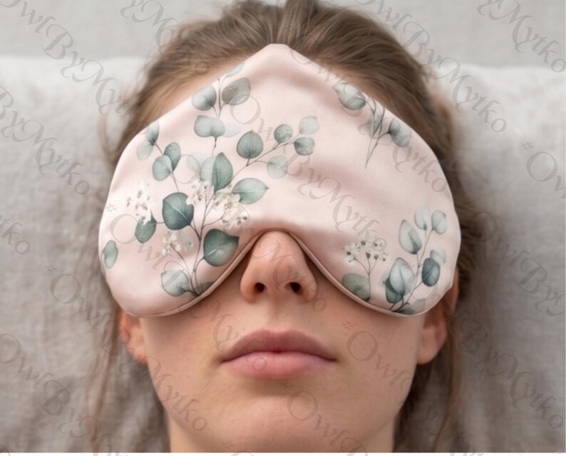 May include: A light pink sleep mask with a botanical design of green eucalyptus leaves and white flowers. The eye mask is made of a soft material and is designed to block out light for better sleep.