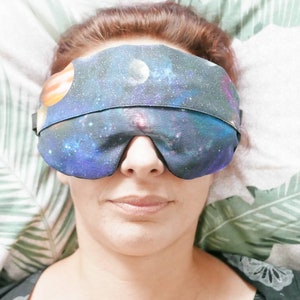 May include: A person wearing a sleep mask with a galaxy print. The mask is blue, purple and black with a white planet and orange ring.