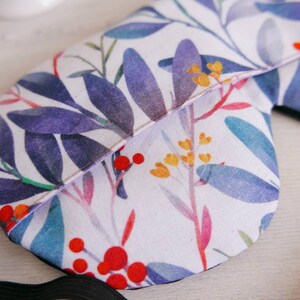 May include: A white sleep mask with a floral pattern featuring blue, purple, red, yellow, and green watercolour leaves and flowers. The mask has a black elastic strap.