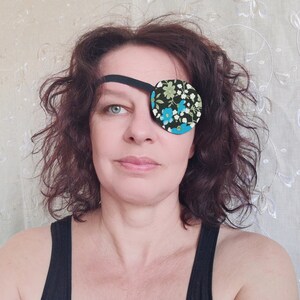 May include: A woman with brown curly hair is wearing a black eye patch with a floral pattern in shades of blue, green, and white. The eye patch is secured with a black elastic band.