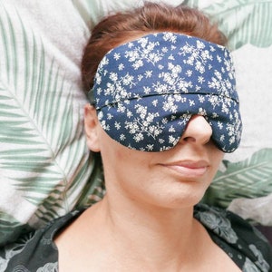 May include: A navy blue sleep mask featuring a white floral design. The mask is made of fabric and has a strap to secure it. Designed to block out light, it aids in better sleep and relaxation.
