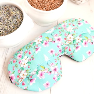 May include: A turquoise eye mask with a floral print of white and pink flowers. The mask is made of fabric and has a black elastic band.