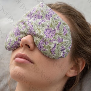 May include: A floral eye mask with a pattern of purple berries and green leaves. The mask is made of soft fabric and is designed to cover the eyes. The background is a light gray.