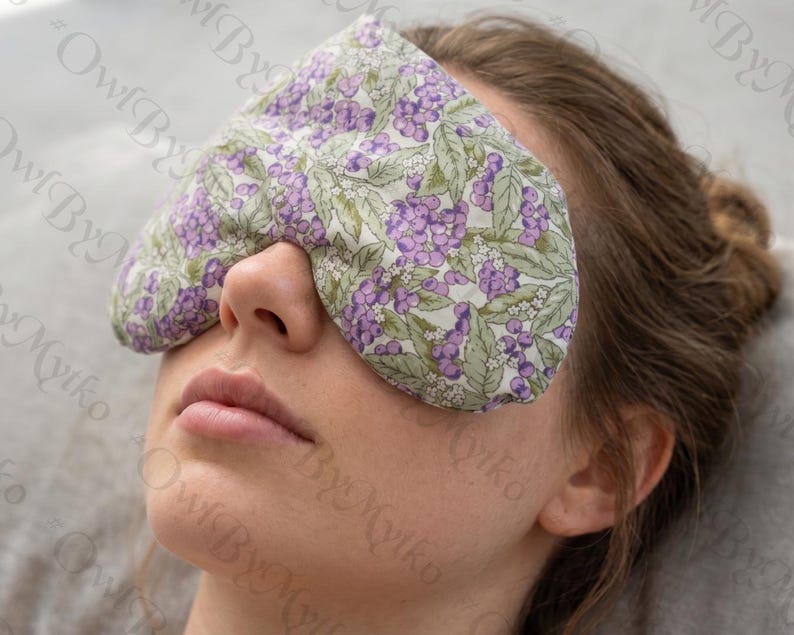May include: A floral eye mask with a pattern of purple berries and green leaves. The mask is made of soft fabric and is designed to cover the eyes. The background is a light gray.