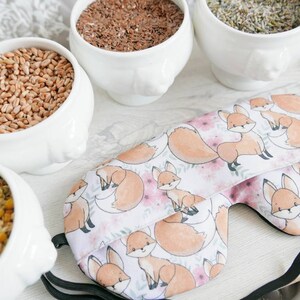May include: A fabric eye mask featuring a repeating pattern of orange foxes and pink flowers on a white background, accompanied by three white ceramic bowls filled with brown grains, dried yellow corn, and dried lavender buds.