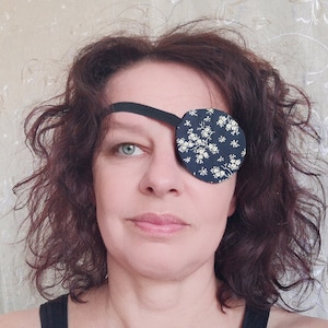 May include: A woman with brown curly hair is wearing a black eye patch with a white floral pattern. The eye patch is secured with a black elastic band.