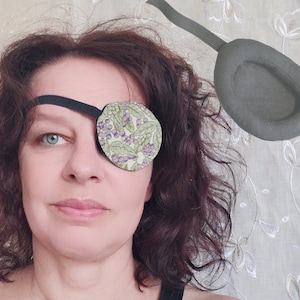 May include: An eye patch featuring a light green fabric with a pattern of purple berries and green leaves, attached to a black elastic strap. A second, solid dark grey eye patch with a matching black elastic strap is shown in the upper right.