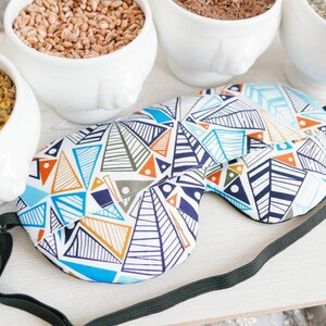 May include: A sleep mask with a geometric pattern in blue, orange, and white. The mask has a black elastic strap. White bowls filled with grains and herbs surround the mask.