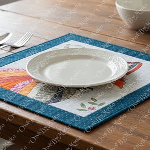 Puede incluir: Un plato de cer&aacute;mica blanca con borde decorativo descansa sobre un mantel individual cuadrado. El mantel individual presenta un dise&ntilde;o de p&aacute;jaro colorido con tonos naranja, azul y verde, enmarcado por un borde azul. Se ve cuberter&iacute;a sobre la mesa de madera.