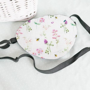 May include: A white eye mask with a floral print featuring pink and purple flowers and small bees. The mask has an adjustable black elastic strap.