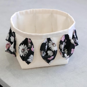 Può includere: Un piccolo cestino in tessuto bianco sporco con un motivo floreale. Il cestino ha una forma rettangolare con angoli arrotondati ed è decorato con foglie di tessuto nero con fiori bianchi, grigi e rosa. L'interno è bianco sporco.