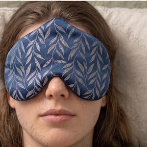 May include: A navy blue sleep mask with a silver leaf pattern. The mask is made of a soft, silky material and is designed to block out light. The sleep mask is a sleep accessory.