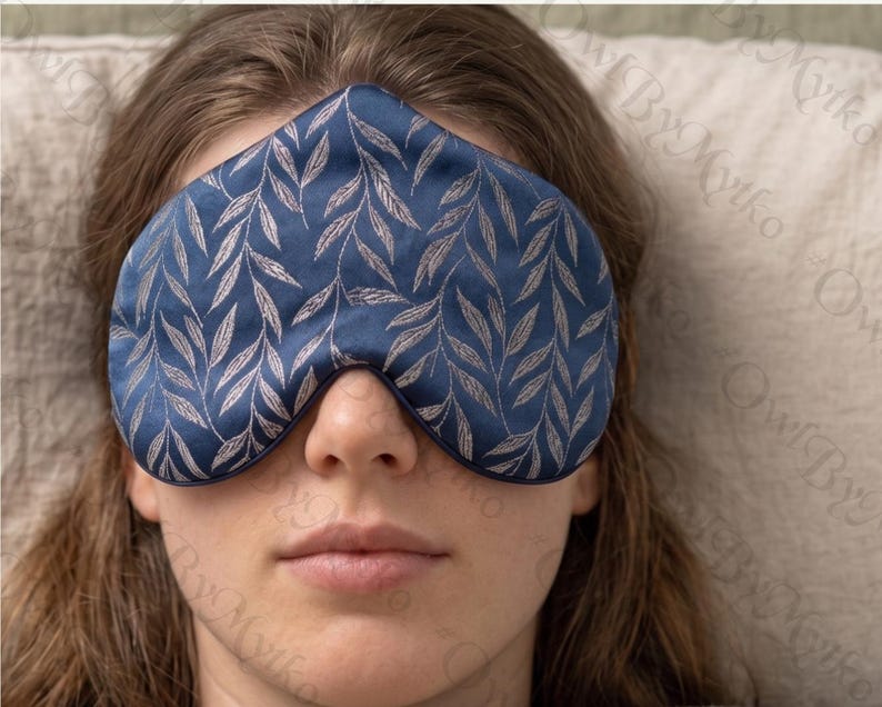 May include: A navy blue sleep mask with a silver leaf pattern. The mask is made of a soft, silky material and is designed to block out light. The sleep mask is a sleep accessory.