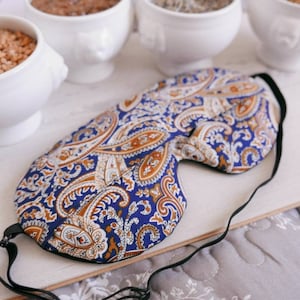 May include: A sleep mask featuring a vibrant blue, white and tan paisley pattern. The mask has a contoured shape with a black elastic strap and adjustable hardware, designed to cover the eyes for light blocking during rest or travel.