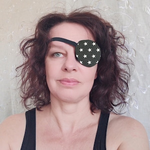 May include: A woman with brown hair is wearing a black eye patch with white stars. The eye patch is secured with a black strap.