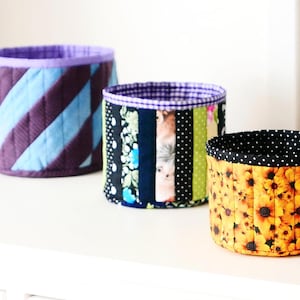 May include: Three fabric storage containers. One is purple and blue striped, another has a patchwork design, and the third features yellow sunflowers on a black background. The containers have a cylindrical shape and are lined with contrasting fabrics.