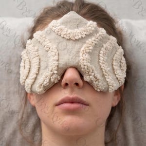 May include: A beige eye mask with a textured, looped design. The mask covers the eyes and nose, with a soft, neutral color palette. The mask is made of a soft material.