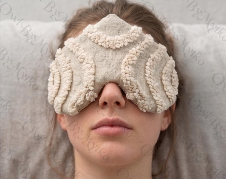 May include: A beige eye mask with a textured, looped design. The mask covers the eyes and nose, with a soft, neutral color palette. The mask is made of a soft material.