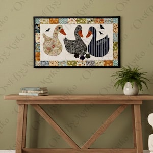 May include: A framed textile art piece featuring three geese with patterned bodies and orange beaks. The artwork is bordered by a patchwork design and hangs above a wooden console table. A small plant in a white pot sits on the table.