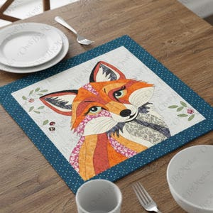 Puede incluir: Un mantel individual cuadrado con un dise&ntilde;o de zorro colorido. El zorro tiene pelaje naranja, rojo y blanco con ojos verdes. El mantel individual tiene un borde azul con lunares blancos y est&aacute; colocado sobre una mesa de madera con un plato, un cuenco y cubiertos.