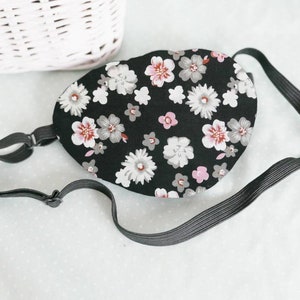 May include: Black sleep mask with a floral pattern in shades of pink, white and grey. The mask has an adjustable elastic strap.