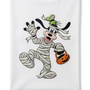 Op de afbeelding: Een wit t-shirt met een geborduurde afbeelding van een stripfiguurhond verkleed als mummie. Het personage draagt lichtbeige windsels, een groene hoed en houdt een oranje pompoen-snoepmandje vast met een zwart uitgesneden gezicht.