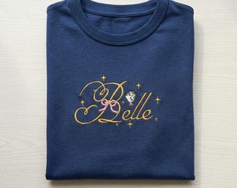 May include: A navy blue t-shirt with the embroidered word "Belle" in gold script, accented with a pink bow and small star details. A small illustration of a character is also embroidered on the shirt.