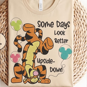 May include: A beige t-shirt featuring an embroidered graphic of Tigger standing upside down. The design includes three Mickey Mouse-shaped balloons in lime green, pink, and teal, with the text "Some Days Look Better Upside-Down" in black.