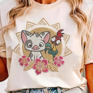 May include: A cream-colored t-shirt featuring an embroidered design of a white piglet with black spots and a red-crested rooster. The characters are centered within a gold sunburst frame, accented by three pink hibiscus flowers and gold leaves.