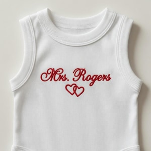 Puede incluir: Body de bebé blanco acanalado con cuello redondo y diseño sin mangas. El body presenta el texto bordado "Mrs. Rogers" en escritura roja, con dos corazones rojos debajo.