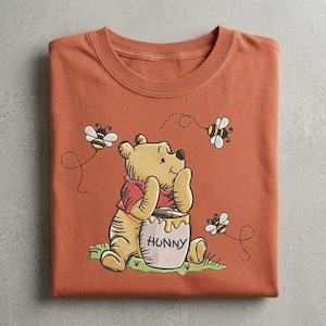 May include: A rust-colored t-shirt featuring an embroidered graphic of a yellow bear wearing a red shirt, sitting behind a honey pot labeled HUNNY. The bear is surrounded by three small flying bees with dashed flight paths on a green patch.