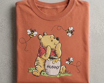 May include: A rust-colored t-shirt featuring an embroidered graphic of a yellow bear wearing a red shirt, sitting behind a honey pot labeled HUNNY. The bear is surrounded by three small flying bees with dashed flight paths on a green patch.